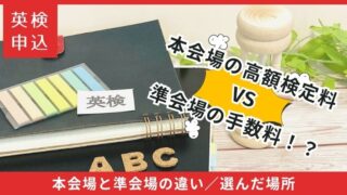 英検申込に関するアイキャッチ画像。黒いノートと付箋、アルファベットオブジェを背景に、「本会場の高額検定料 VS 準会場の手数料！？」という大きな対比文字が配置されている。下部には「本会場と準会場の違い／選んだ場所」と記載。